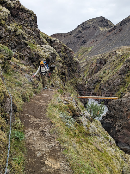 A person walking on a narrow path with a bridge over a canyon in the background.